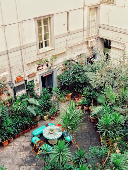 A narrow cobbled pathway in a small courtyard garden features a variety of potted plants and flowers alongside a textured stone wall. The pots are made of different materials, including white ceramic