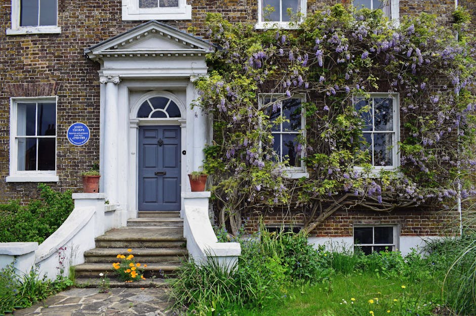 A wooden front door set within a stone porch opening, surrounded by two large, lush, green hedges with dense, glossy leaves. The hedges are neatly trimmed and frame the door symmetrically, creating a 